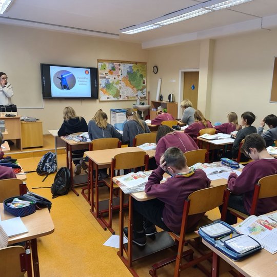 Klasėje, pilnoje mokinių, vyksta istorijos pamoka. Priekyje klasės stovi edukatorė.