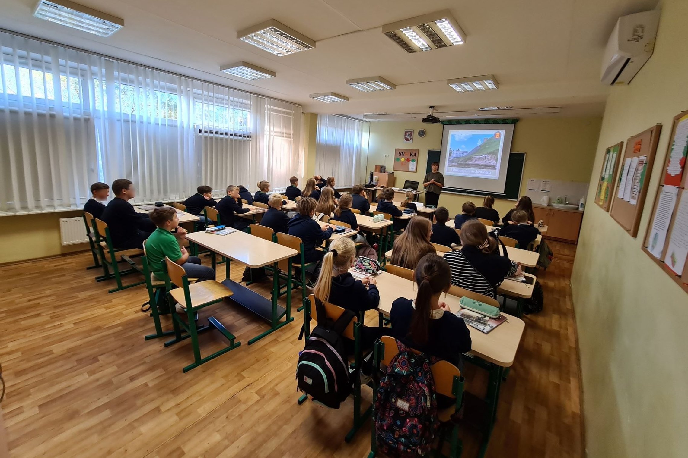 Nuotraukoje vaizduojami mokiniai klasėje. Gale stovi mokytoja ir dėsto temą iš skaidrių, kurios yra pavaizduotos ant projektoriaus ekrano.