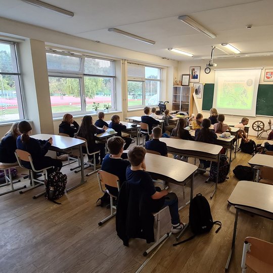 Vaizduojami klasėje sėdintys mokiniai, pro klasės langus matosi mokyklos sporto aikštelė. Klasei yra rodomos skaidrės apie Vilniaus piliakalnius, mokiniai susidomėję žiūri į skaidres.