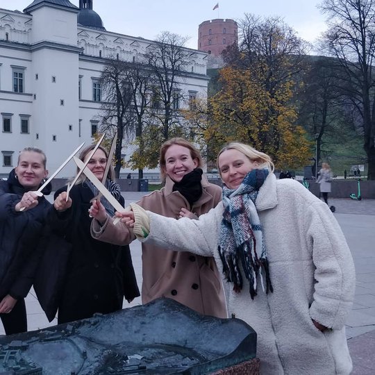Muzikos leidyklos DAMN GOOD prodiuserės šalia Vilniau pilių rezervato maketo Katedros aikštėje. Merginos laiko medinius kalavijus, sudėjusios centre. 