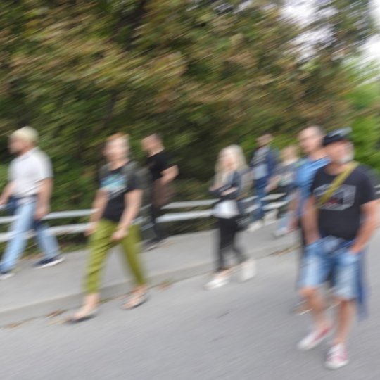 Nuotrauka yra nesufokusuota. Matomi tik žmonių siluetai, kurie eina keliu palei Vilnelę. Upę ir žmones skiria medžiai bei tvorelė.