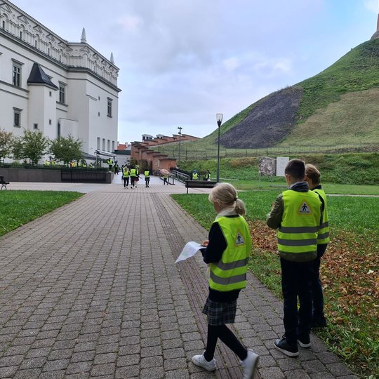 Nuotraukoje trys mokiniai. Gale stovintys du berniukai yra nusisukę ir žiūri į apačią, šalia jų eina mergaitė, kuri rankose laiko žemėlapį. Visa likusi klasė eina priekyje. Nuotraukos kairėje pusėje matosi Valdovų rūmai, o dešinėje - Gedimino pilis.