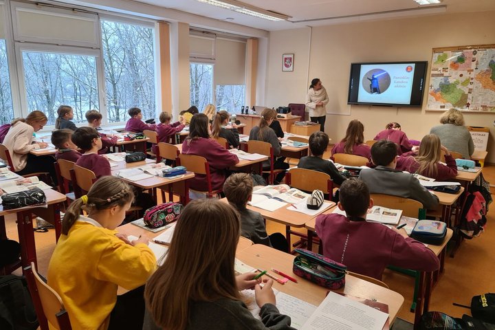 Klasėje, pilnoje mokinių, vyksta istorijos pamoka. Priekyje klasės stovi edukatorė.