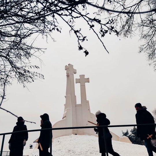 Trijų Kryžių paminklas iš apačios. Priešais keturių praeinančių žmonių siluetai. 