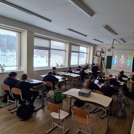 Vaikai klasėje fotografuoti iš nugarų. Projektoriuje matosi puošnūs herbai.