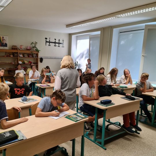 Nuotraukoje vaizduojami progimnazijos mokiniai, sėdintys klasėje. Tarp mokinių suolų stovi mokytoja, kuri kalba apie tos dienos temą.