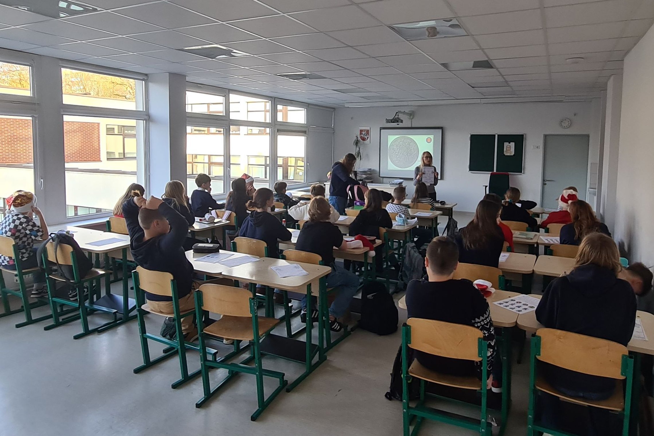 Mokiniai klasės suoluose, fotografuoti iš galo. Priekyje klasės stovi mokytoja.
