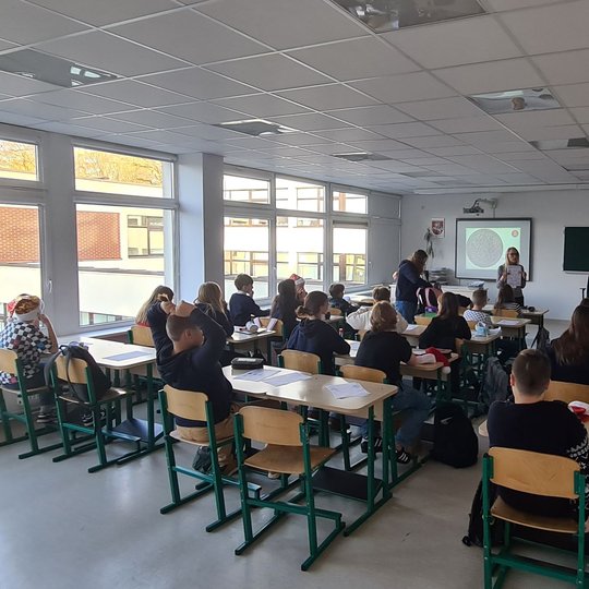 Mokiniai klasės suoluose, fotografuoti iš galo. Priekyje klasės stovi mokytoja.
