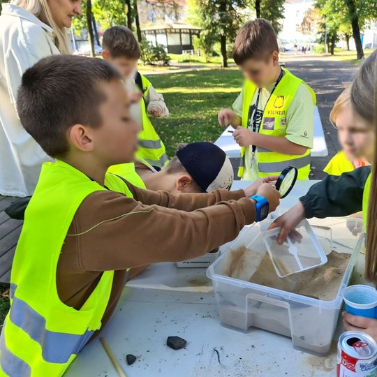 Nuotraukoje matosi, kaip vaikai, apstoję stalą, vykdo mini archeologinius tyrimus, mokosi kaip tai reikia daryti. Ant stalo matosi dėžė su smėliu, vienas iš vaikų laiko lupą, fone matosi Valdovų rūmų parkelis.