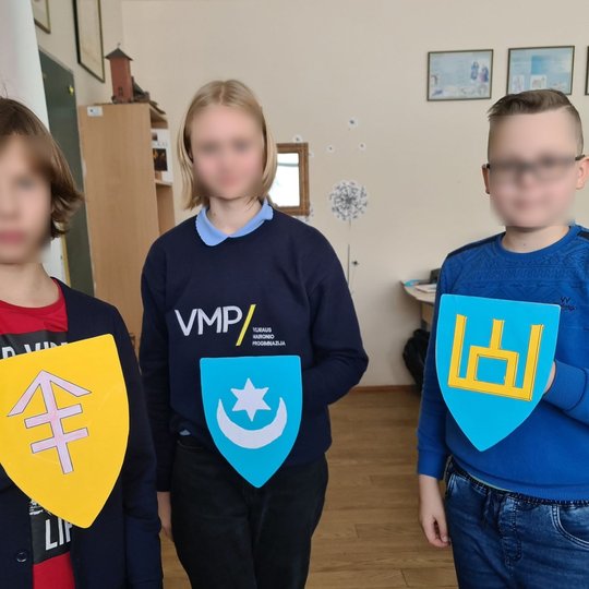 Nuotraukoje trys moksleiviai laiko savo pagamintus spalvingus herbus.