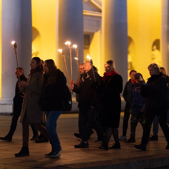 Ekskursantai su fakelais eina per Katedros aikštę, aplink tamsu.