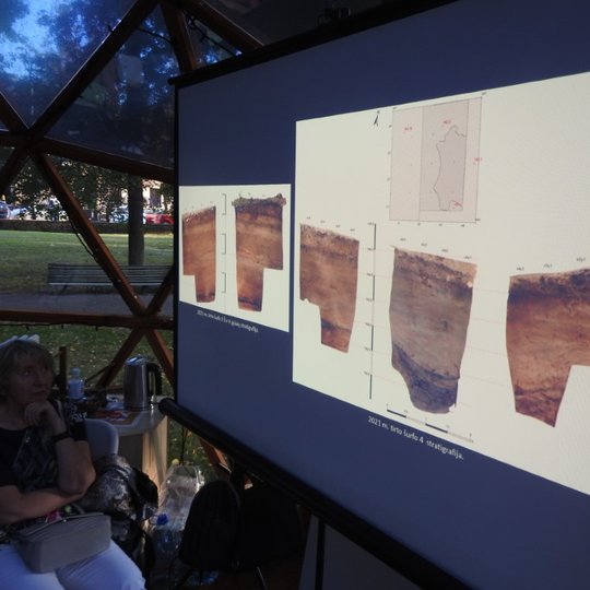 Garbaus amžiaus moteris stebi pateiktą skaidrę apie archeologinius radinius. Skaidrėje matomi penki žemės sluoksnių pavyzdžiai.
