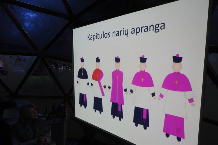 Nuotrauka vaizduoja skaidrę, kuri yra ant projektoriaus ekrano. Skaidrėje yra skirtingos kapitulos narių aprangos. Šios aprangos yra pavaizduotos piešinio formatu ir virš jų yra parašytas skaidrės pavadinimas ryškai juodomis raidėmis "Kapitulos narių apra