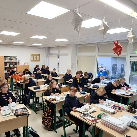 Nuotraukoje vaizduoja klasėje prie savo suolų sėdinčius penktokus. Kai kurie šypsosi kamerai, o kiti rašo savo sąsiuviniuose. Nuotrauka padaryta iš klasės priekio.