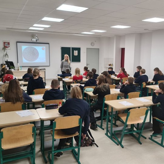 Mokiniai klasės suoluose, fotografuoti iš galo. Priekyje klasės stovi mokytoja.