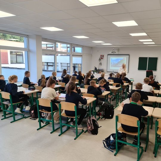 Nuotrauka padaryta iš klasės galo. Klasėje trisdešimt mokinių. Mokytoja stovi priekyje ir aiškina temą, skaitydama vadovėlį. Atrodo, kad tema įdomi, nes visi vaikai klauso susikaupę.