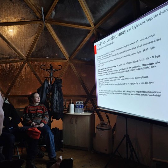 Vilniaus pilių stikliniame kupole stovi projektoriaus ekranas prie kurio sėdi žmonės ant kėdžių ir klausosi paskaitos.