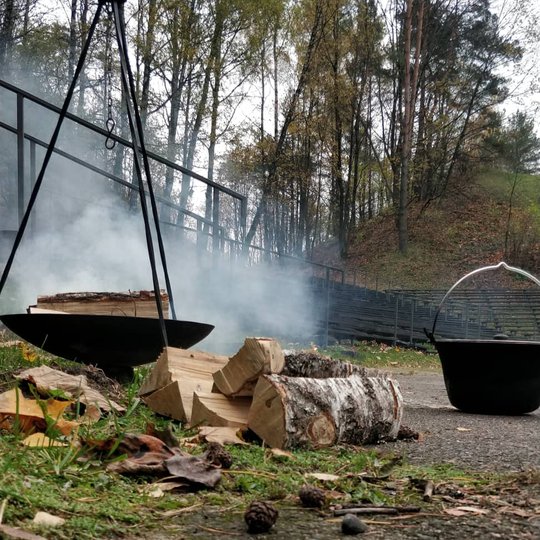 Rūkstantis laužas su katilu ir malkomis šalia.