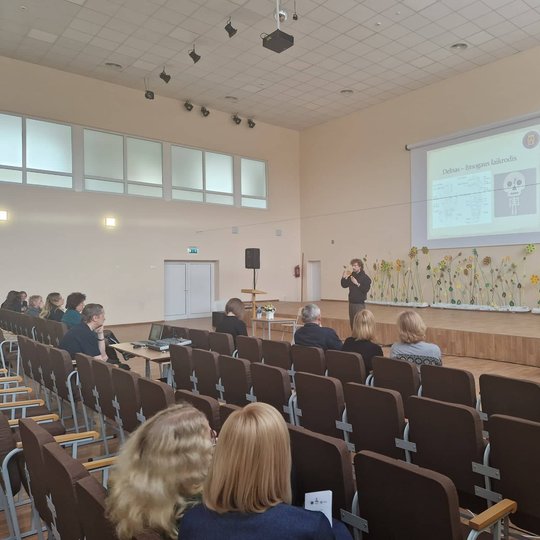 Edukatorius Vytas Navickas gimnazijos aktų salėje skaito pranešimą. Jam už nugaros ekranas su skaidrėmis.