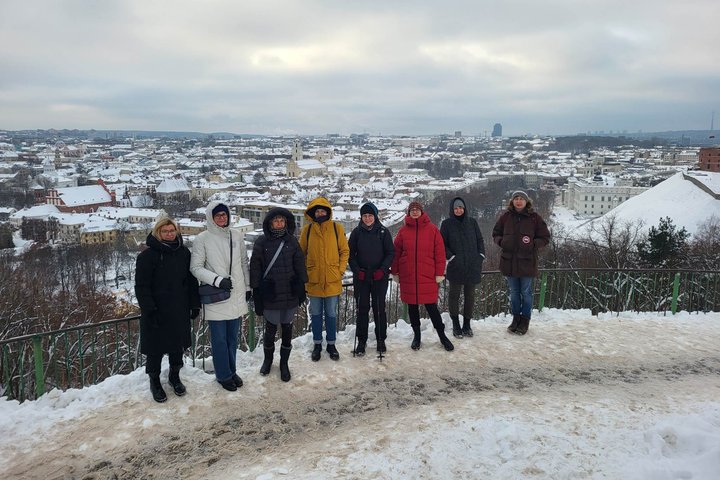 Ekskursijos dalyviai pozuoja nuotraukai nuo Trijų Kryžių kalvos. Jiems už nugaros apsnigto ir žiemiško Vilniaus miesto ir senamiesčio  panorama.