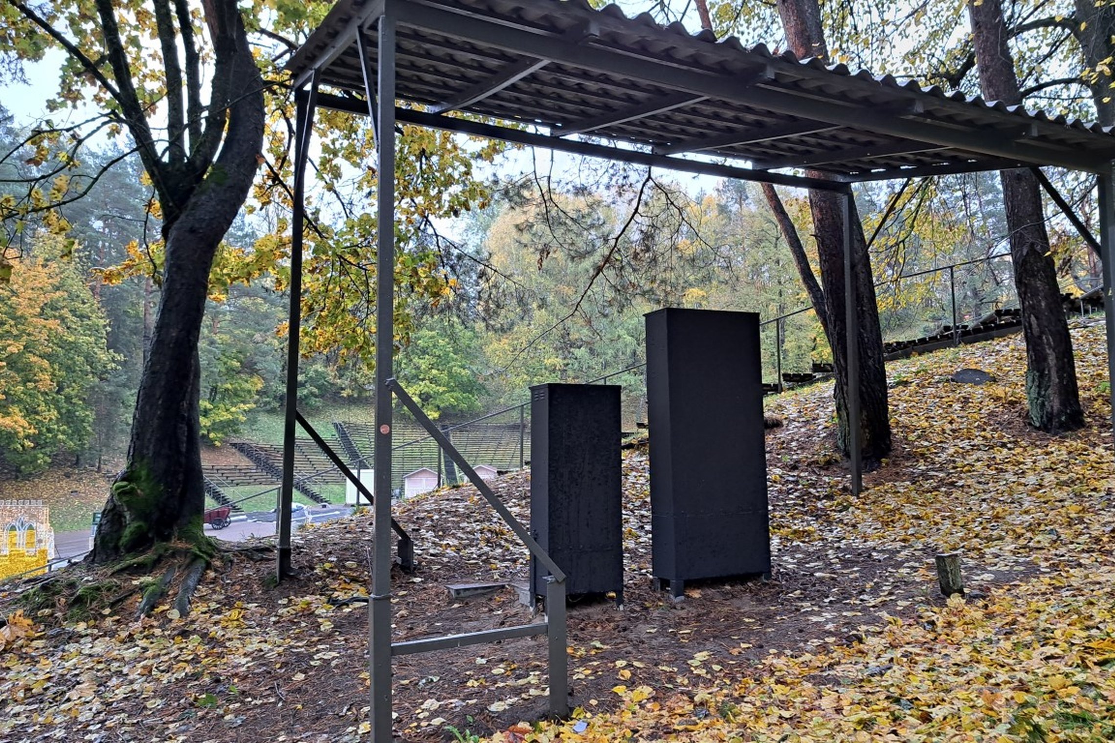 Nuotrauka daryta prie Kalnų parko estrados. Joje stovi du elektros skydeliai, vienas didesnis, kitas mažesnis, virš jų pastatytas stogelis. Taip pat matosi daug, nuo medžių, prikritusių, geltonų lapų, todėl vaizdas atrodo labai rudeninis.