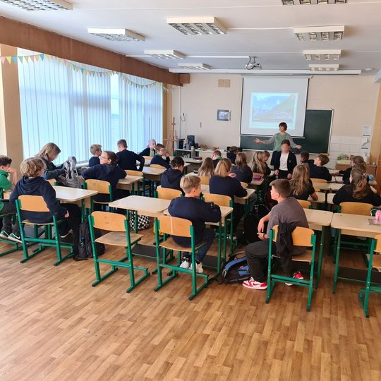 Klasės priekyje stovinti mokytoja mokiniams rodo skaidres. Mokiniai klausosi.