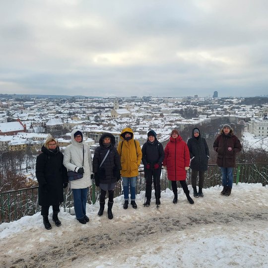 Ekskursijos dalyviai pozuoja nuotraukai nuo Trijų Kryžių kalvos. Jiems už nugaros apsnigto ir žiemiško Vilniaus miesto ir senamiesčio  panorama.