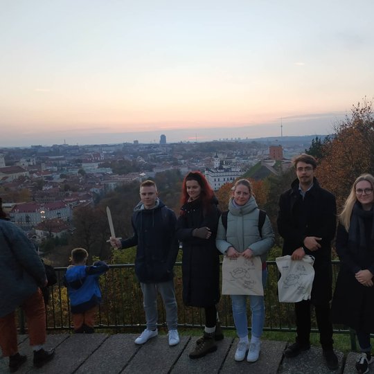 Draugai ir kolegos pozuoja nuotraukai priešais Trijų Kryžių paminklą. Už nugarų Gedimino kalnas su pilimi.