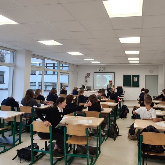 Mokiniai klasės suoluose, fotografuoti iš galo. Priekyje klasės stovi mokytoja.