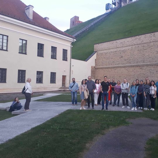 Žmonių grupė fotografuojasi, taip pat už jų matomas muziejaus pastatas ir maža dalis Gedimino pilies.