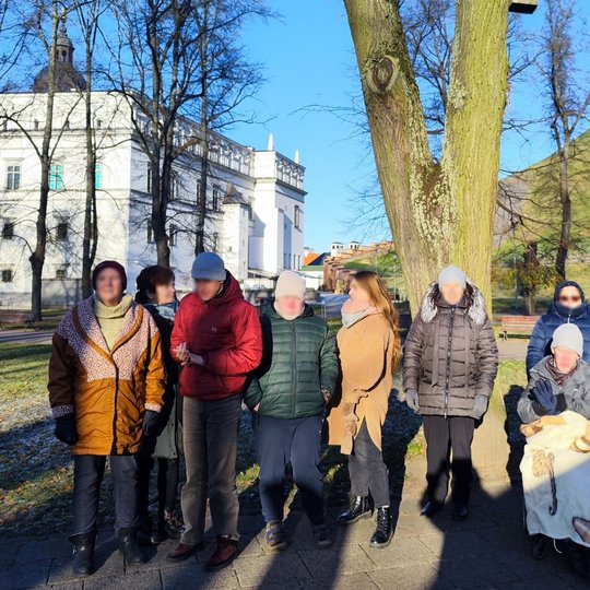 Grupė žmonių su negalia pozuoja Pilių parke, tarp Valdovų rūmų ir Gedimino pilies kalno. Į juos šviečia saulė.