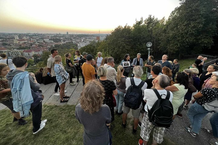 Nuotrauka padaryta ant Trijų kryžių kalno. Vyksta ekskursija. Žmonės sustoję ratu aplink gidę, kuri veda ekskursiją ir laiko rankoje nuotrauką. Turistai labai įdėmiai klauso.