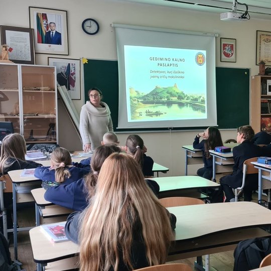 Penktokai žiūri į projektoriaus ekraną ir edukatorę.