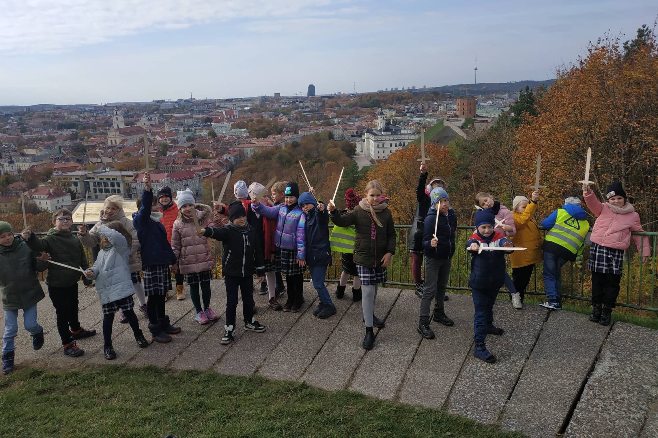 Antrokai pozuoja nuotraukai su mediniais kalavijais ant Trijų Kryžių kalvos. Už nugarų matosi Gedimino kalnas su pilimi.