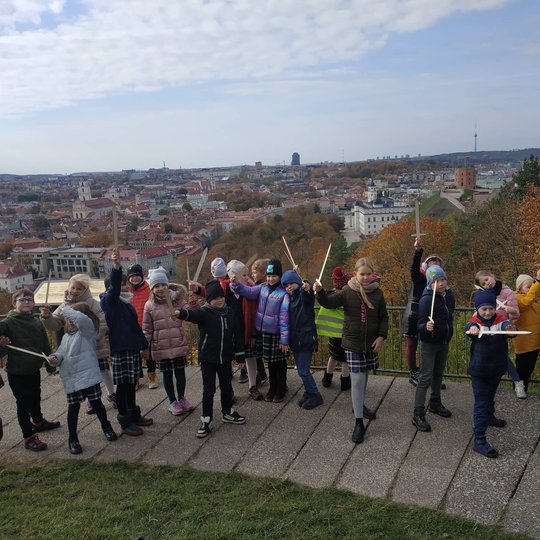 Antrokai pozuoja nuotraukai su mediniais kalavijais ant Trijų Kryžių kalvos. Už nugarų matosi Gedimino kalnas su pilimi.