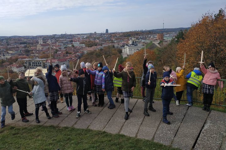 Antrokai pozuoja nuotraukai su mediniais kalavijais ant Trijų Kryžių kalvos. Už nugarų matosi Gedimino kalnas su pilimi.