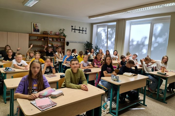 Klasėje sėdintys vaikai šypsosi nuotraukai. Dalis vaikų yra iškėlę nykštį į viršų, kiti sėdi ramiai, linksmai nusiteikę. Iš vadovėlių galime matyti, jog moksleiviai sėdi istorijos kabinete.