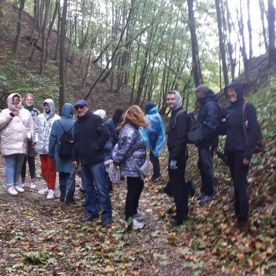 Nuotrauka vaizduoja grupę žmonių, kurie stovi tarp medžių. Jie dėvi striukes ir paltus, šypsosi nuotraukai. Aplinkoje matosi daug medžių, kurie yra jau numetę savo lapus ,o žemė yra pilna šių nukritusių spalvotų lapų.