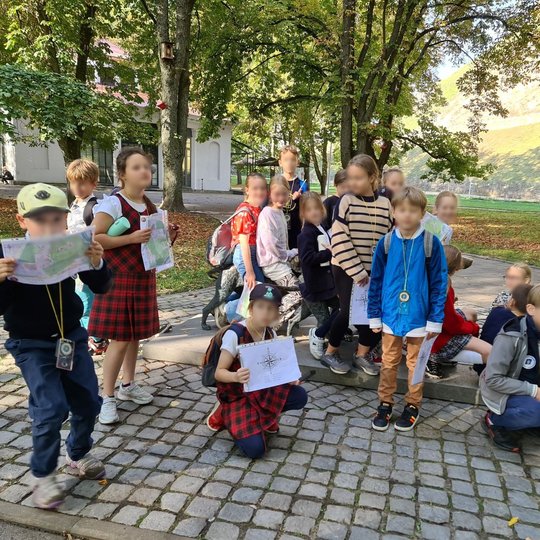 Nuotrauka vaizduoja parke esančius pradinukus. Jie visi yra sustoję šalia vienos kito, pozuoja nuotraukai. Kiekvienas laiko po žemėlapį ir ant kaklo yra užsidėję po kompasą. Už jų matosi daug žalių medžių.