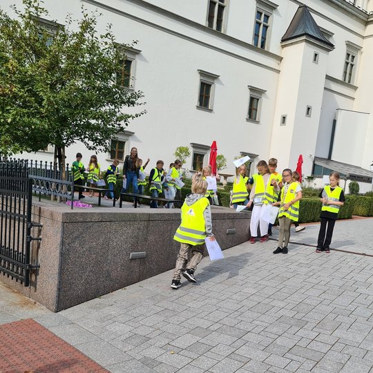 Nuotraukoje grupė trečiokų su ryškiomis žaliai geltonomis liemenėmis. Nuotrauka padaryta, kai vaikai juda ir žiūri į įvairias puses. Vieni stovi, kiti eina ir žvalgosi, o keli bėgioja, vienintelė jų mokytoja iš tolo žiūri į kamerą ir šypsosi. Vaikai stovi