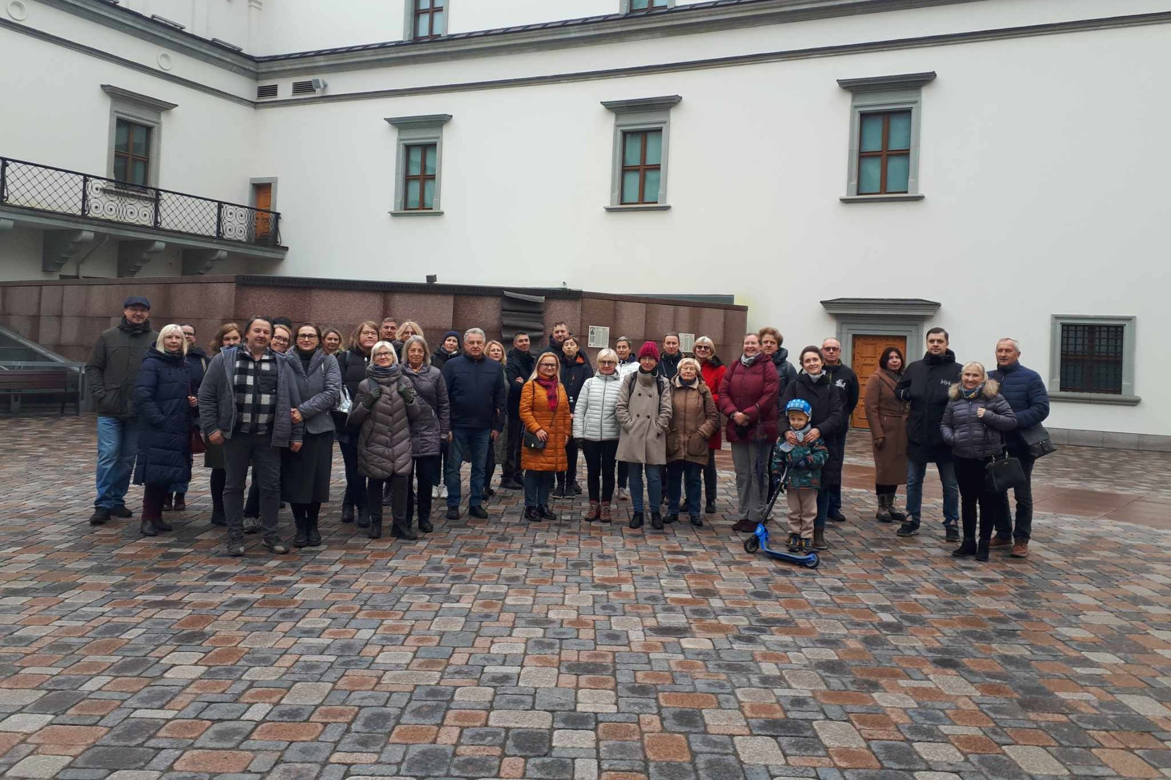 Nuotraukoje vaizduojama didelė grupelė įvairaus amžiaus žmonių. Dauguma žmonių apsirengę šiltus drabužius dėl vėsaus oro. Už žmonių galime matyti Valdovų rūmus, grupelė stovi pastato kieme.