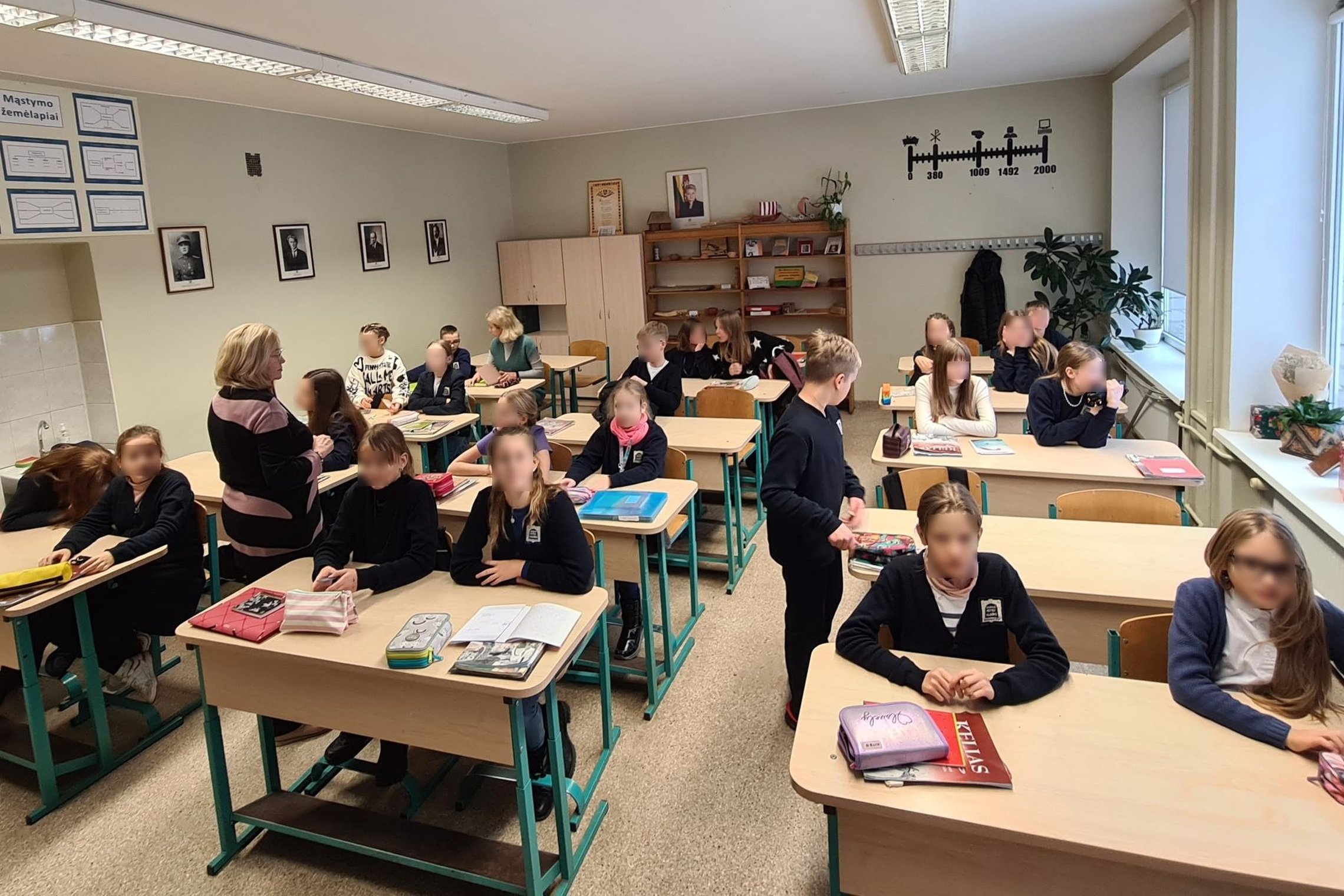 Vaikai ruošiasi istorijos pamokai klasėje. Tarp jų vaikšto ir mokytoja.