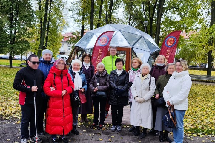 Nuotraukoje matomi žmonės, stovintys prie kupolo, kuris priklauso Vilniaus pilims. Už jų taip pat yra matomi medžiai, suolai. Nuotraukos kairėje pusėje, žmonių puslankio krašte, stovi vyras su lazdele.