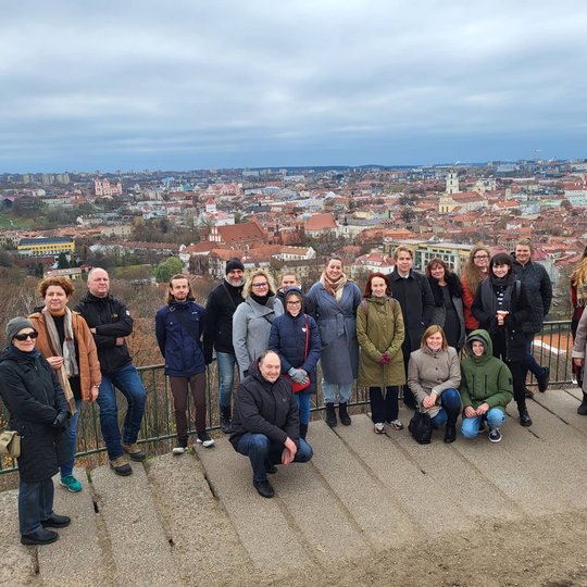 Ekskursijos dalyviai pozuoja nuotraukai priešais Trys Kryžius. Tolumoje matosi Vilniaus panorama.