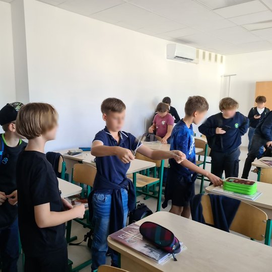 Tarp suolų stovi grupelė penktokų. Vienas iš jų žaidžia su saga ant virvutės, kiti du stebi jį. Kiek toliau nuo jų stovintys vaikai žiūri į savas puses. Ant suolų sudėti vadovėliai.