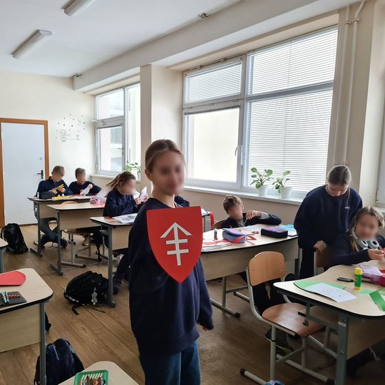 Vaikas klasės viduryje, tarp suolų, rodo savo pagamintą herbą.
