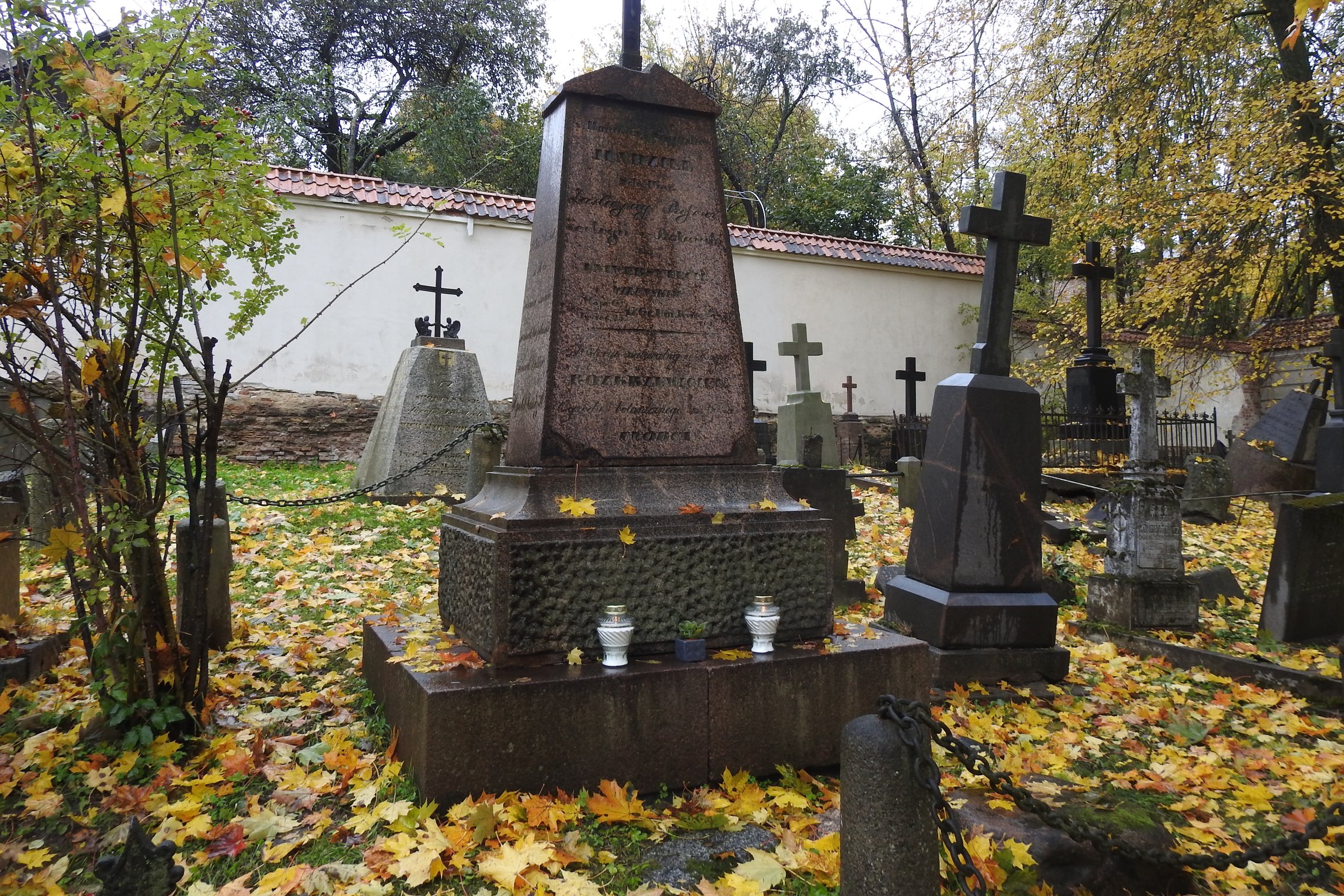 Nuotraukos centre - Jundzilų kapas. Ant antkapio krašto padėtos dvi žvakės, o tarp jų mažas augalėlis vazonėlyje. Šone ir užnugaryje stovi daugiau antkapių, kai kur kapus skiria grandinės. Už betoninės sienos auga medžiai.