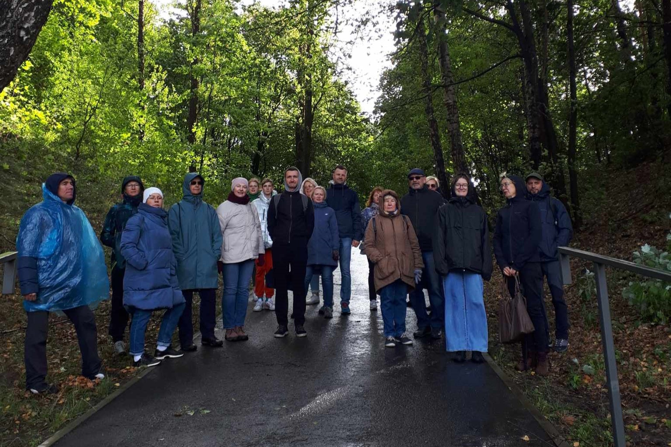 Nuotraukoje matome, kaip ant pėsčiųjų tako, apaugusio tankiu mišku, stovi grupelė žmonių. Žmonės dėvi rudeninio sezono striukes.