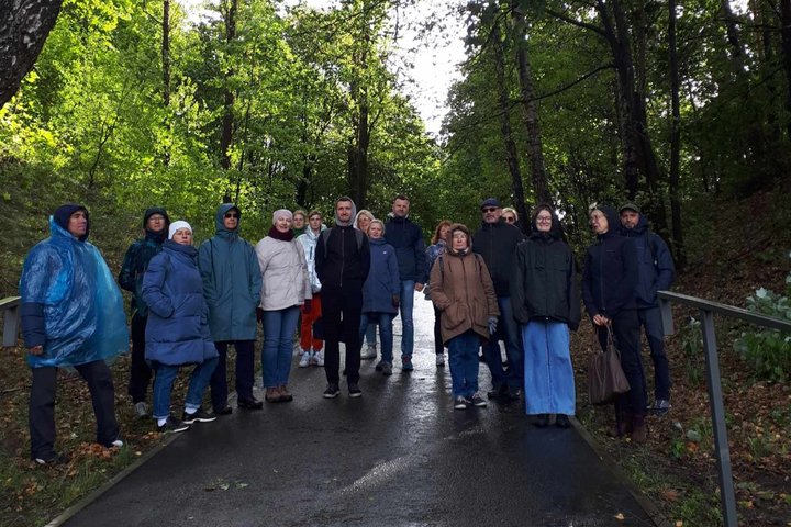 Nuotraukoje matome, kaip ant pėsčiųjų tako, apaugusio tankiu mišku, stovi grupelė žmonių. Žmonės dėvi rudeninio sezono striukes.