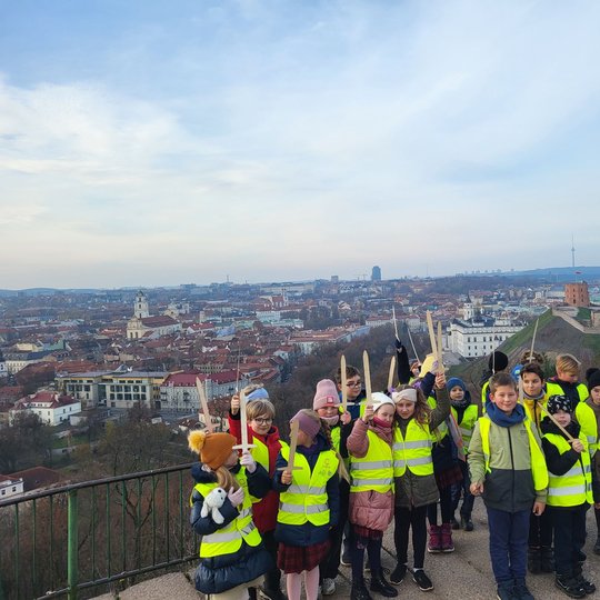 Mokiniai, su atšvaitinėmis liemenėmis, pozuoja nuotraukai  su mediniai kalavijai rankose ant Trijų Kryžių kalvos. Tolumoje matosi Vilniaus panorama.